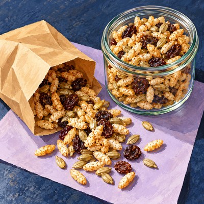 Cinnamon-coated puffed rice, raisins and sunflower kernels spill from a paper snack bag beside a clear jar.